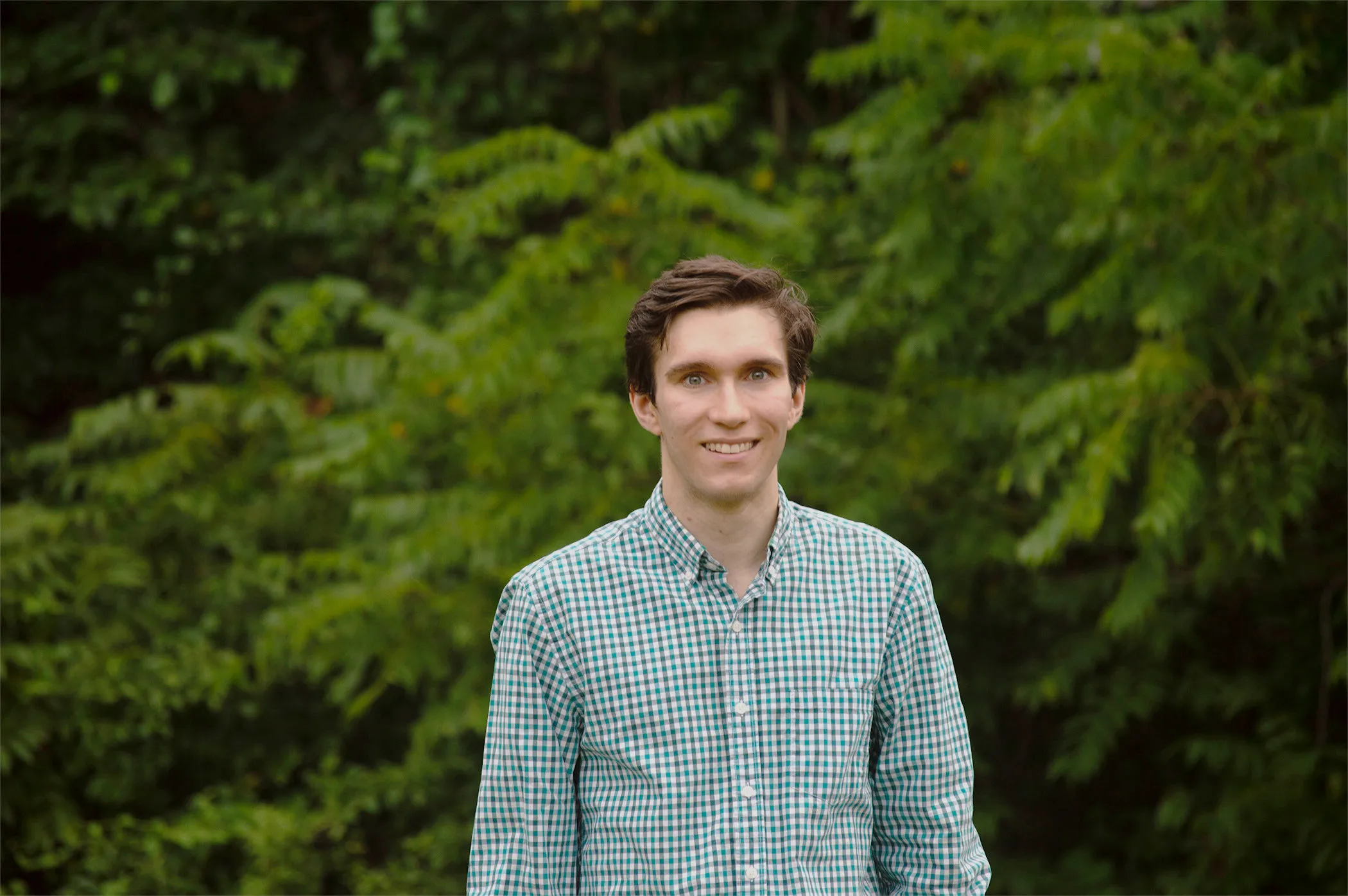 A photo portrait of Spencer Ainsworth, cropped to show his head and torso. He is smiling, has short, dark hair, and is wearing a light, watch-pattered, button-down shirt with an open collar. In the background, out-of-focus trees and shrubs are visible.