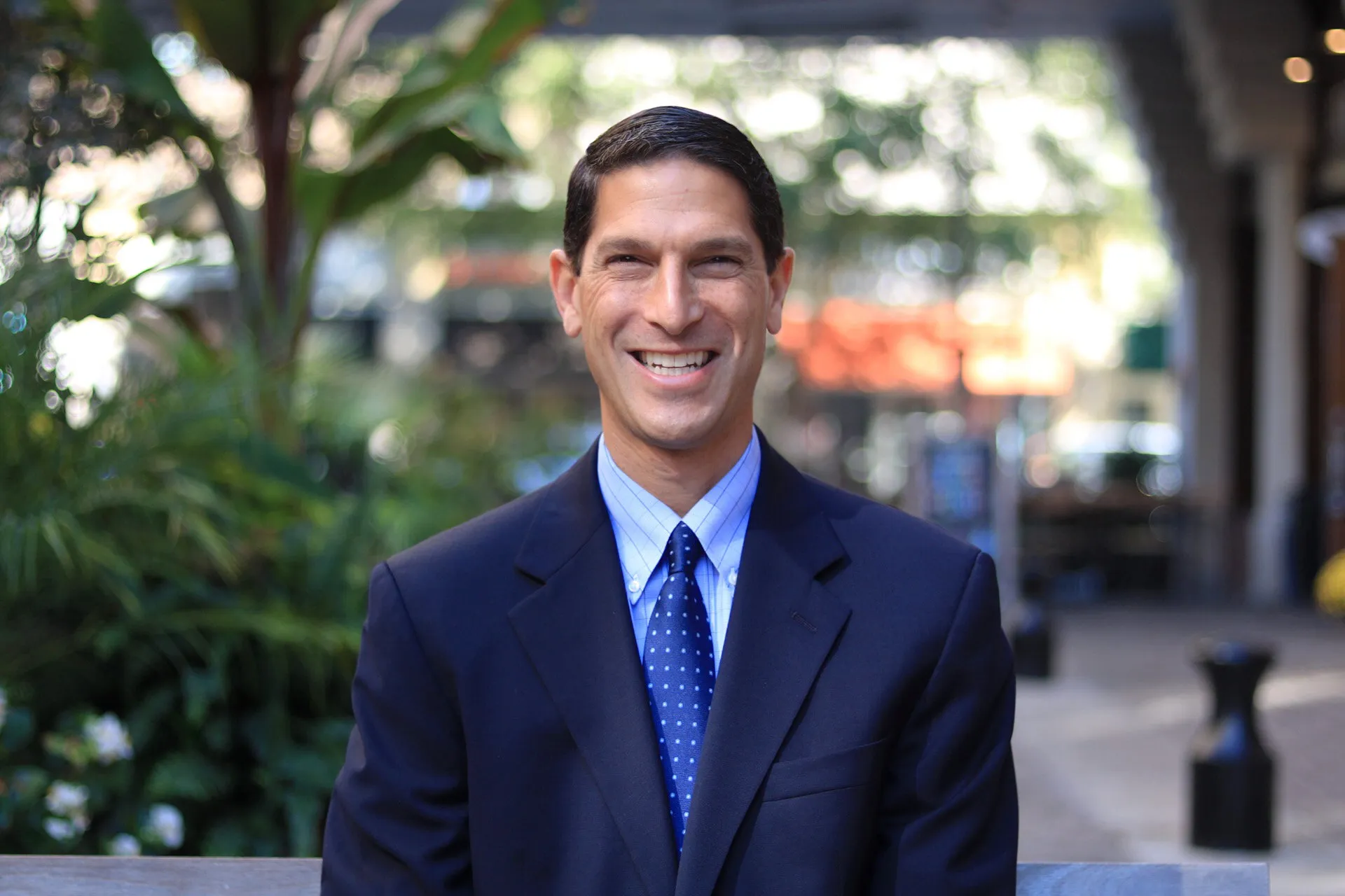 A photo portrait of Joel Eisenfeld, cropped to show his head and upper torso. He is smiling, has short, dark hair, and is wearing a dark suit and necktie. In the background, out-of-focus trees and shrubs are visible.