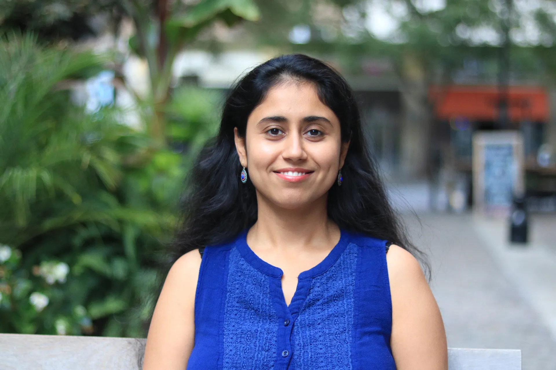 A photo portrait of Ridhima Mehrotra, cropped to show her head and shoulders. She is smiling, has long hair, and is wearing earrings and a sleeveless shirt. In the background, out-of-focus trees and shrubs are visible.