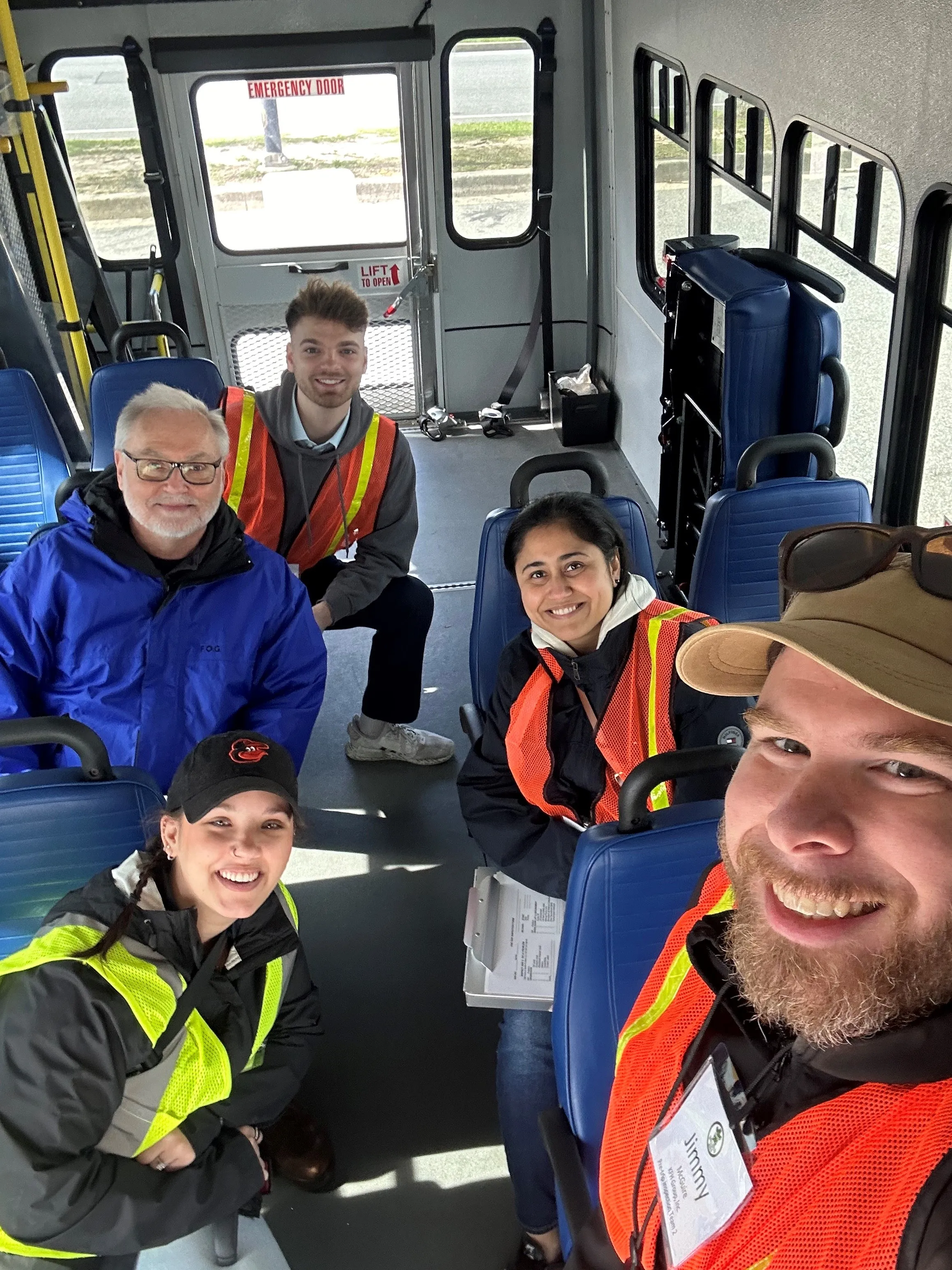 Photo: A group of KFH staff members are seated on a bus at a project site. A few are wearing reflective safety vests and nametags, and carrying clipboards. The point of view is overhead, shot from a camera held at arms' length by Jimmy.