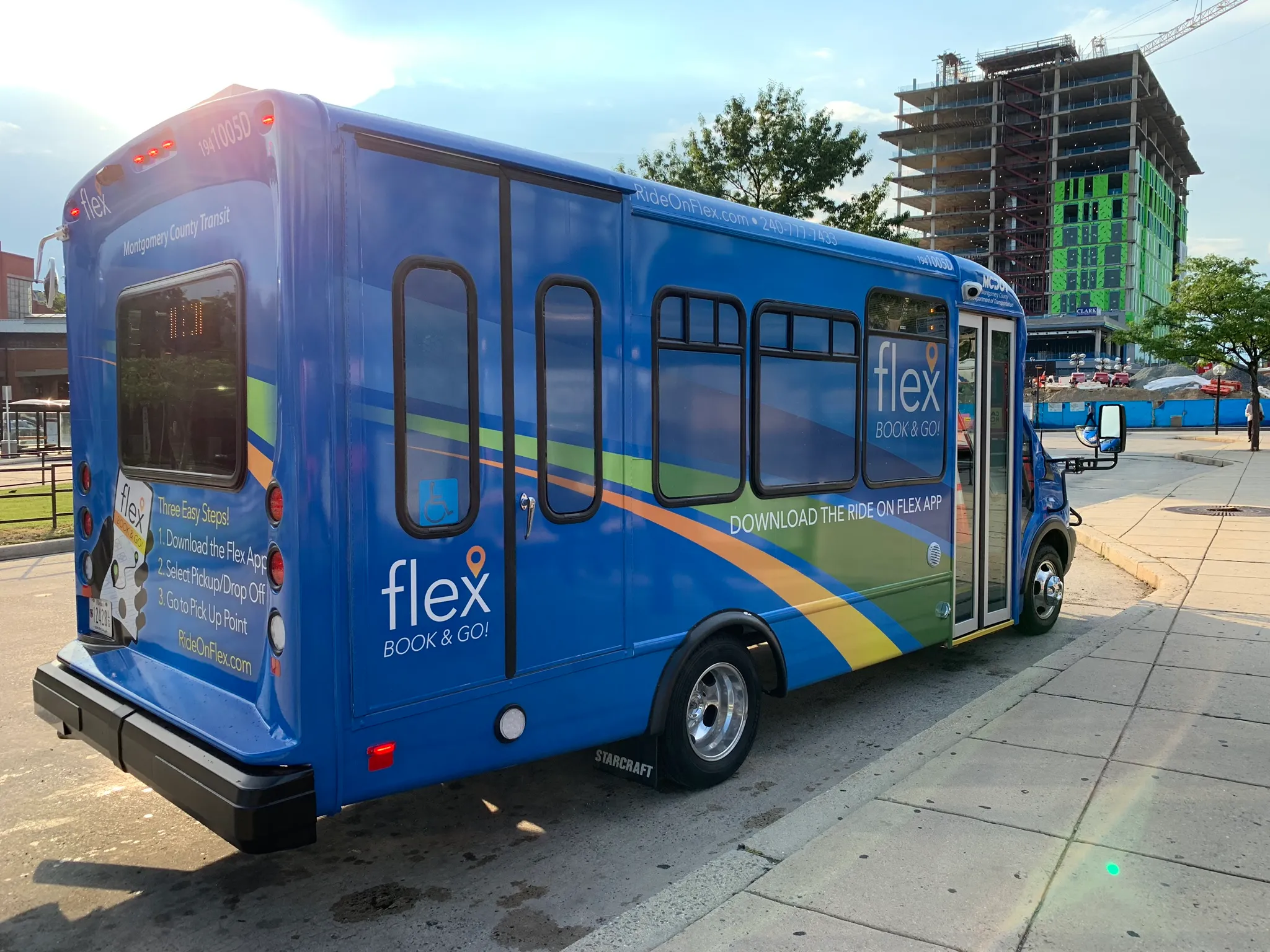 A photo of a Flex Microtransit bus at a bus stop in Montgomery County. The bus, shown at a rear three-quarters angle, is blue with rainbow-stripe graphics on its side.