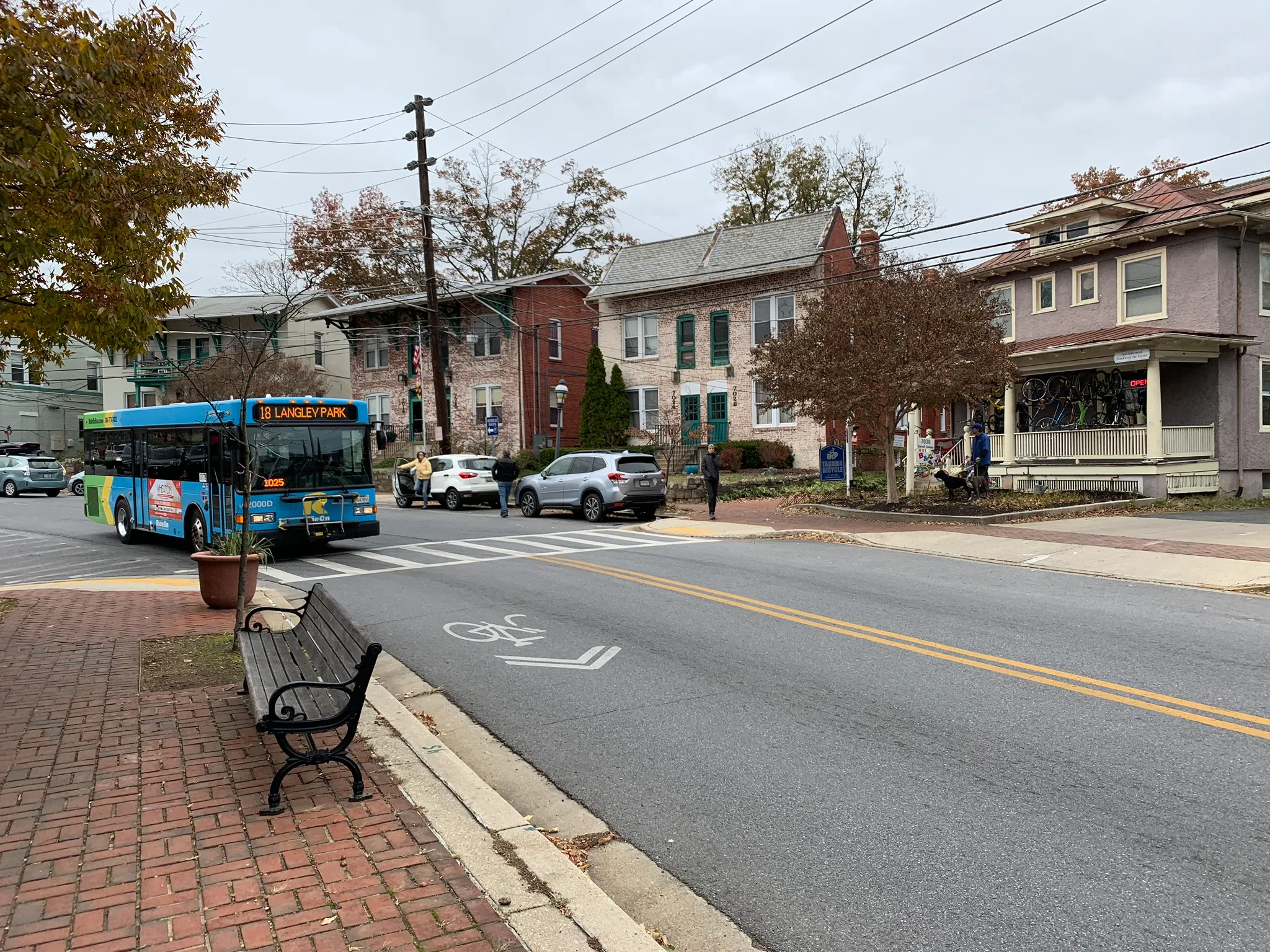 A photo of a bus approaching a stop in Takoma Park, Maryland. The bus is rounding a gentle bend in the road alongside a brick sidewalk. In the road, a striped crosswalk and bicycle right-of-way marking are visible. The bus is bright blue with green and yellow vertical chevron-shaped stripes at the rear.